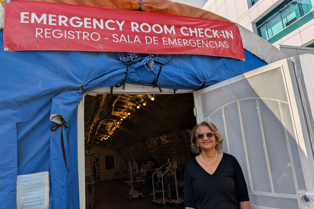 A woman in business formal attire stands beside an entrance to an emergency room check-in.
