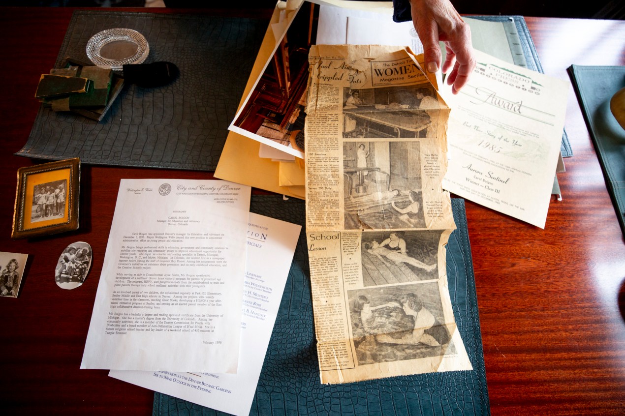Several pieces of paper are arranged on a table. One is a professional biography of Carol Boigon from the Denver City Council. Next is a clipping from The Detroit Times. Last is a 1985 Colorado Press Award.
