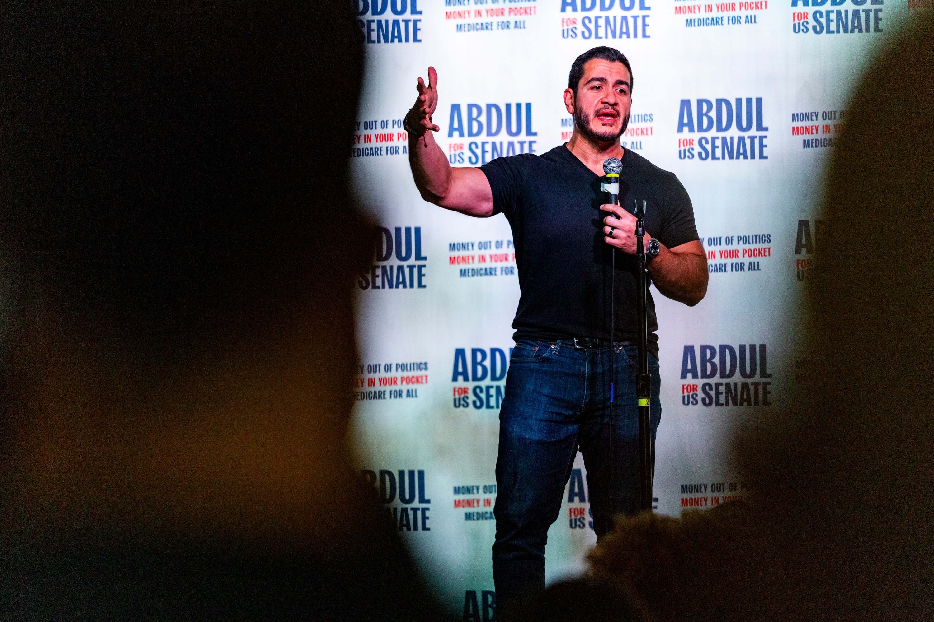 Abdul El-Sayed speaks on-stage at a venue. Behind him is a backdrop that reads, "Abdul for US Senate." Blurred in the foreground are the silhouettes of two heads of people watching El-Sayed speak.