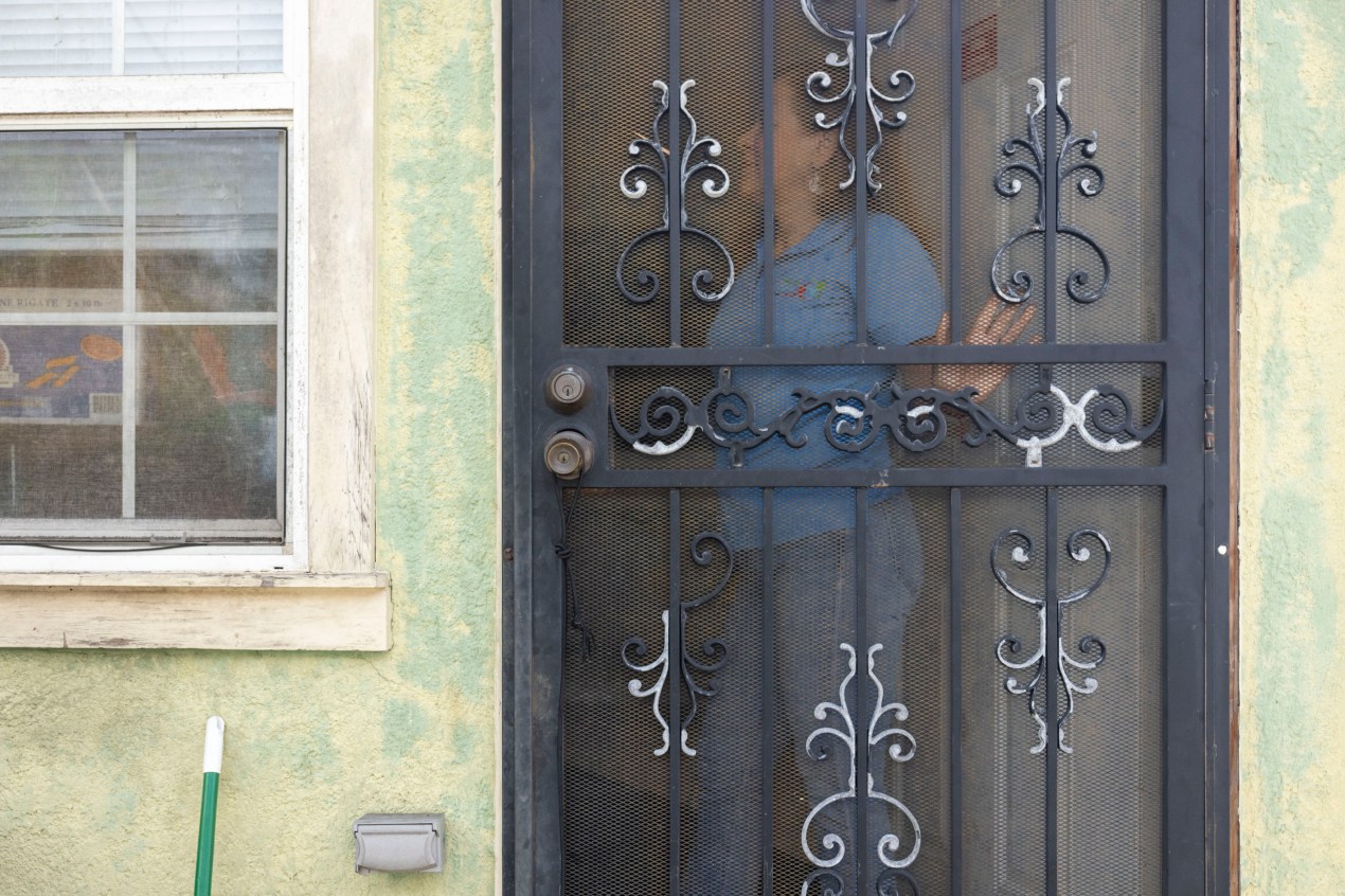 A view from outside looking into a building through a door with screen where a woman stands with her hand to the door as if she's about to push it open.