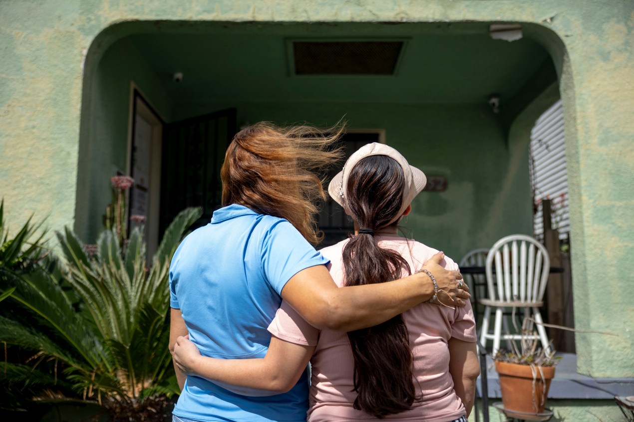 Two women stand side-by-side, each with an arm around the other, and face away from the camera toward a building.