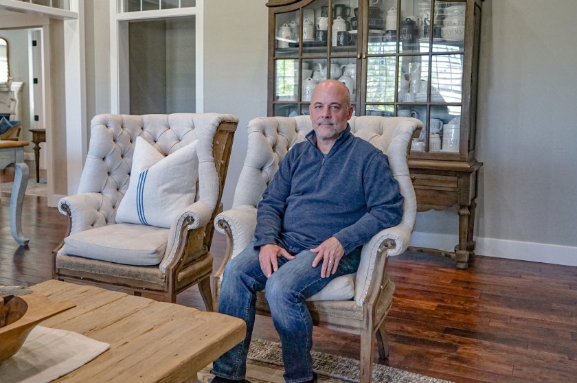 A man in half-zip fleece and jeans since in a light gray armchair and looks at the camera.