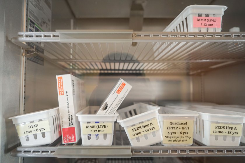 A close-up image through a glass door of the inside a refrigerator where white plastic containers with labels such as "MMR (LIVE) greater than or equal to 12 mo" are lined up on shelves.