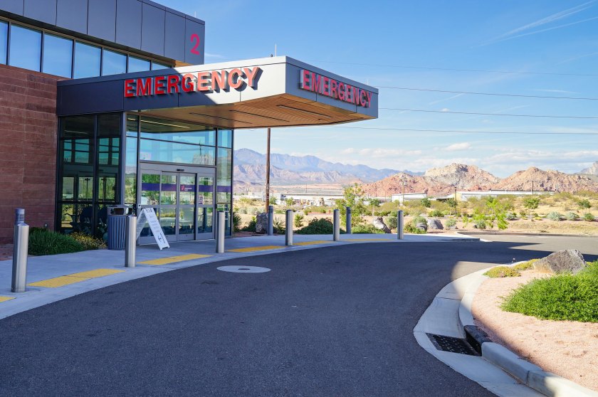 The emergency department entrance to a hospital is shown in the foreground and mountains are visible in the distance behind it.