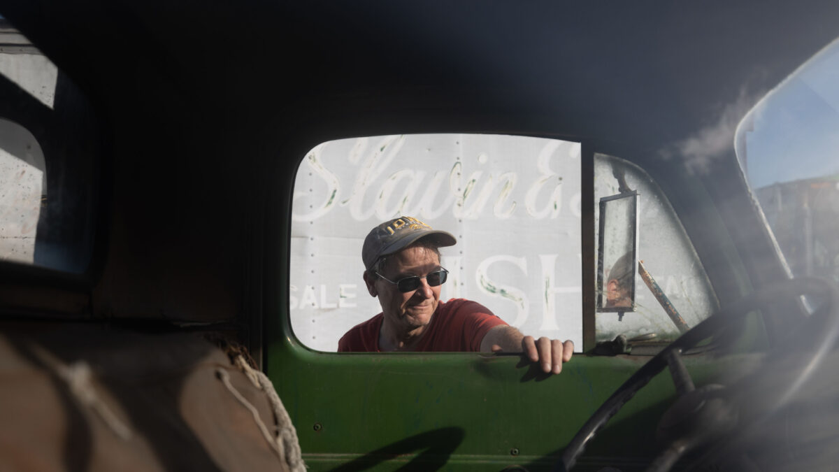 A man wearing a red shirt, a baseball cap and sunglasses rests his hand on the open window of a truck