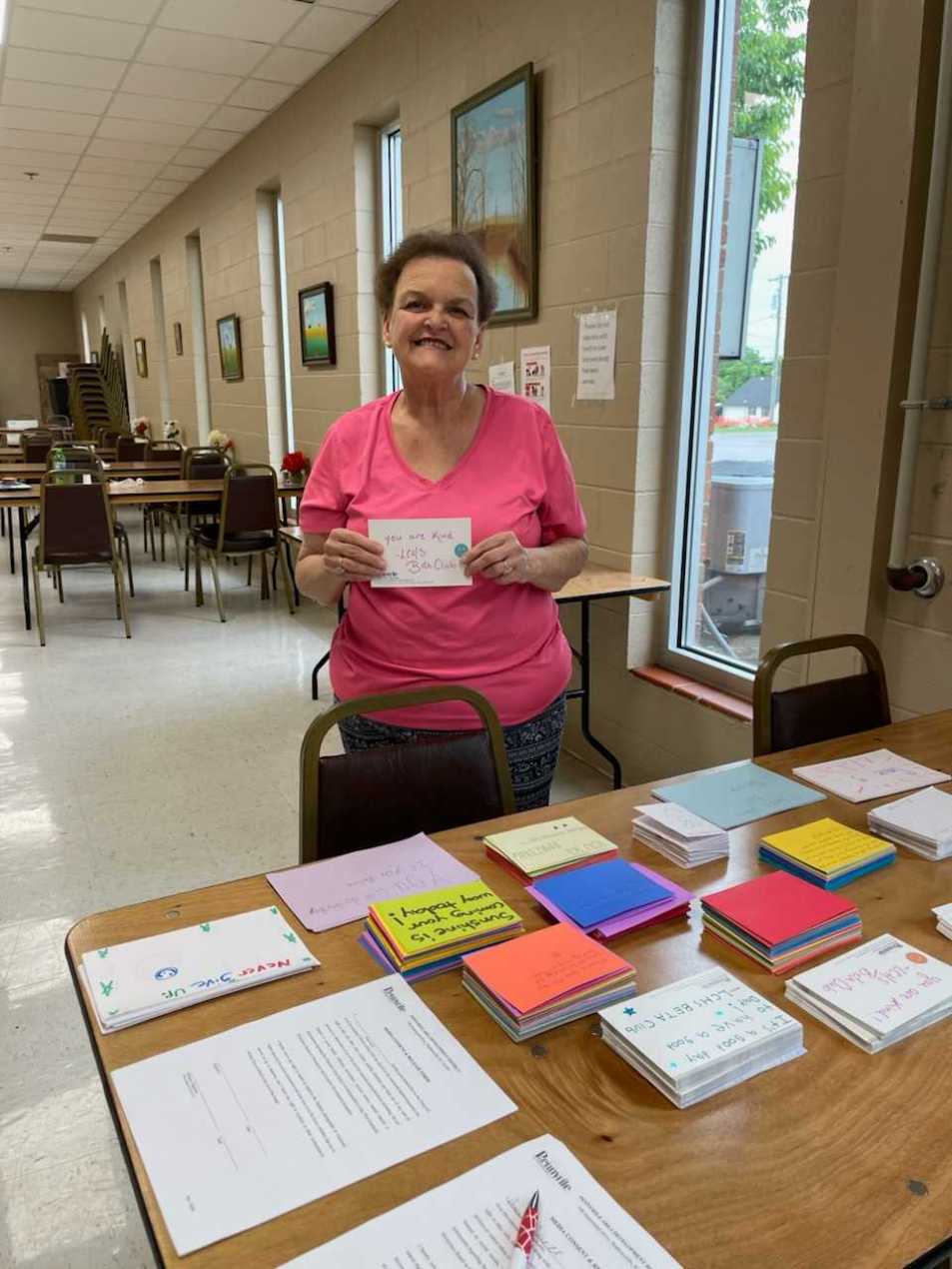 A woman wearing a pink v-neck shirt smiles and holds up a card that reads "you are kind" as she stands in front of a table