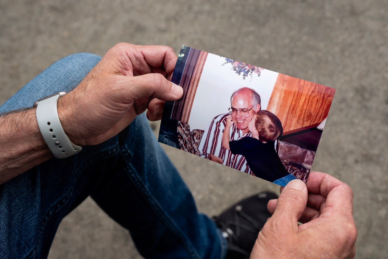 Two hands hold a photo of an older man wearing a striped shirt and glasses who is being hugged and kissed on the cheek by a small boy