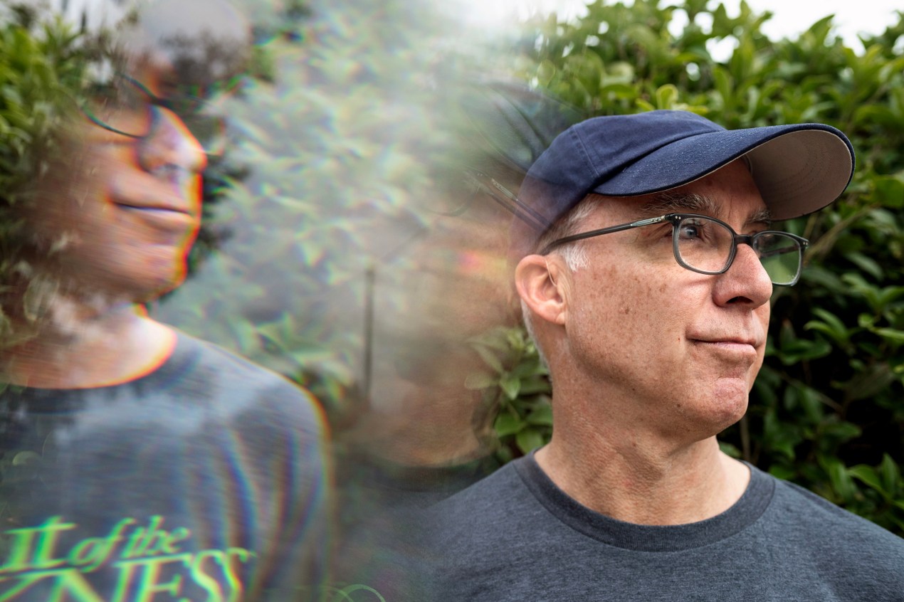 A man wearing a navy baseball cap and glasses stands in front of a green bush and looks off to the side of the frame