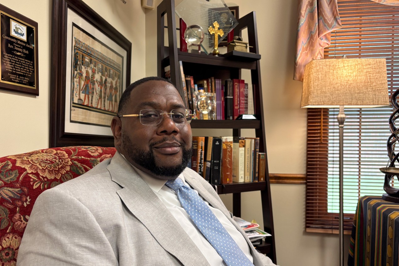 A Black man sits in an interior room. A placard, art print, and bookshelf are seen behind him.