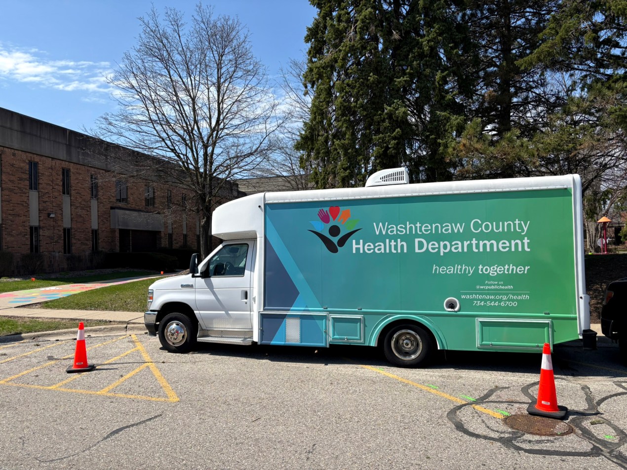 A van with signage that says "Washtenaw County Health Department" is parked in a parking lot next to two orange traffic cones