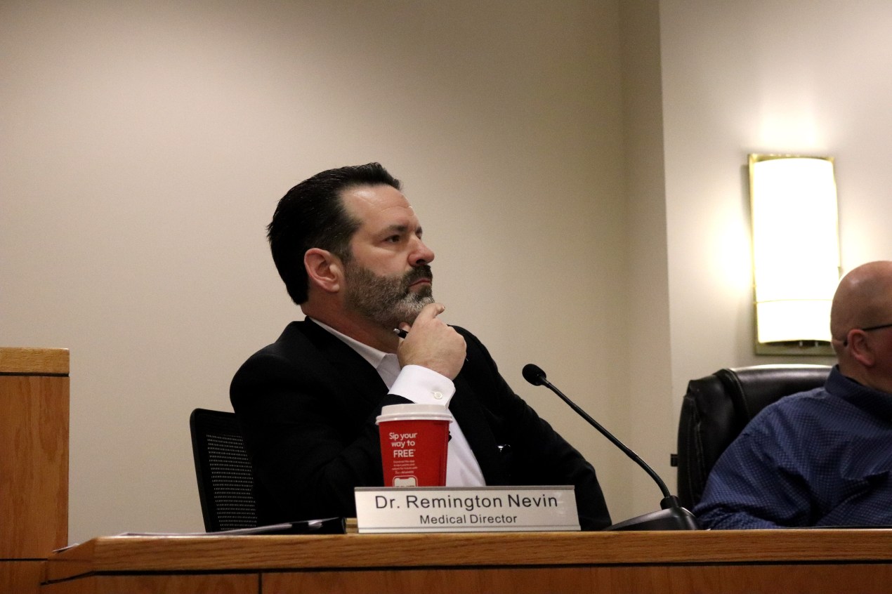 A man sits in front of a microphone with a name placard in front of him that reads "Dr. Remington Nevin, Medical Director"